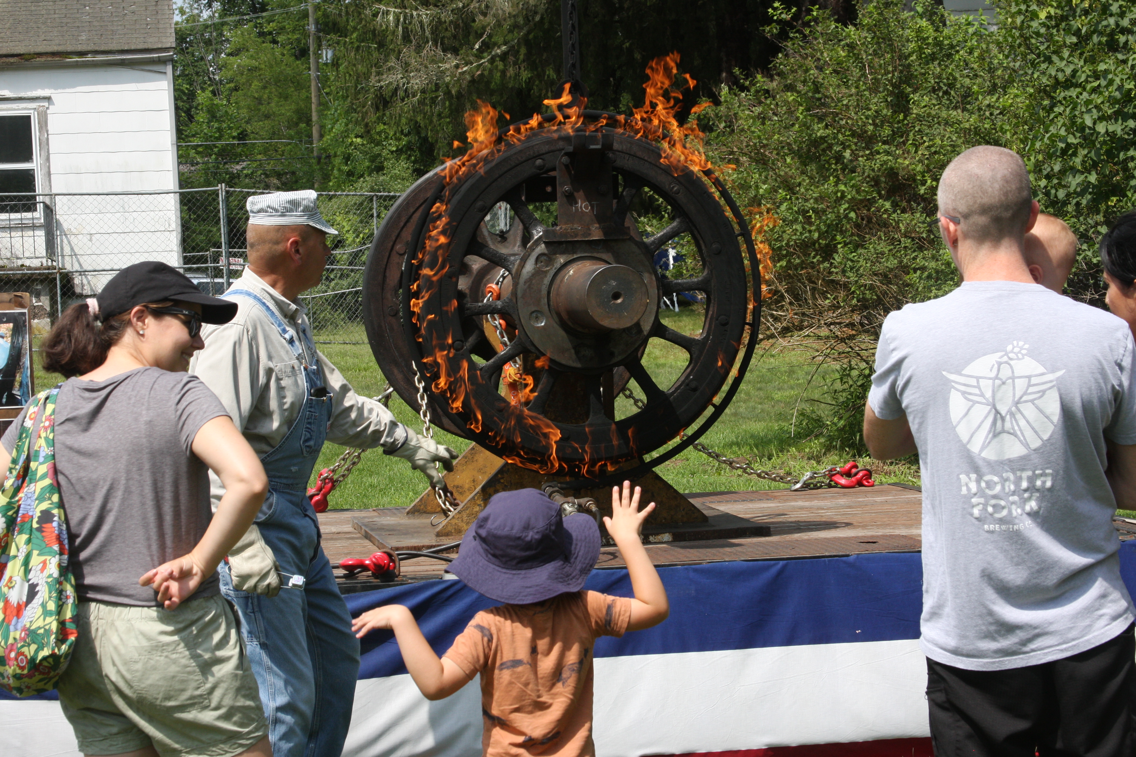 A family watch as man in train engineer clothes pulls on metal chain attached to large metal wheel. The wheel is one fire and mounted on a stand.