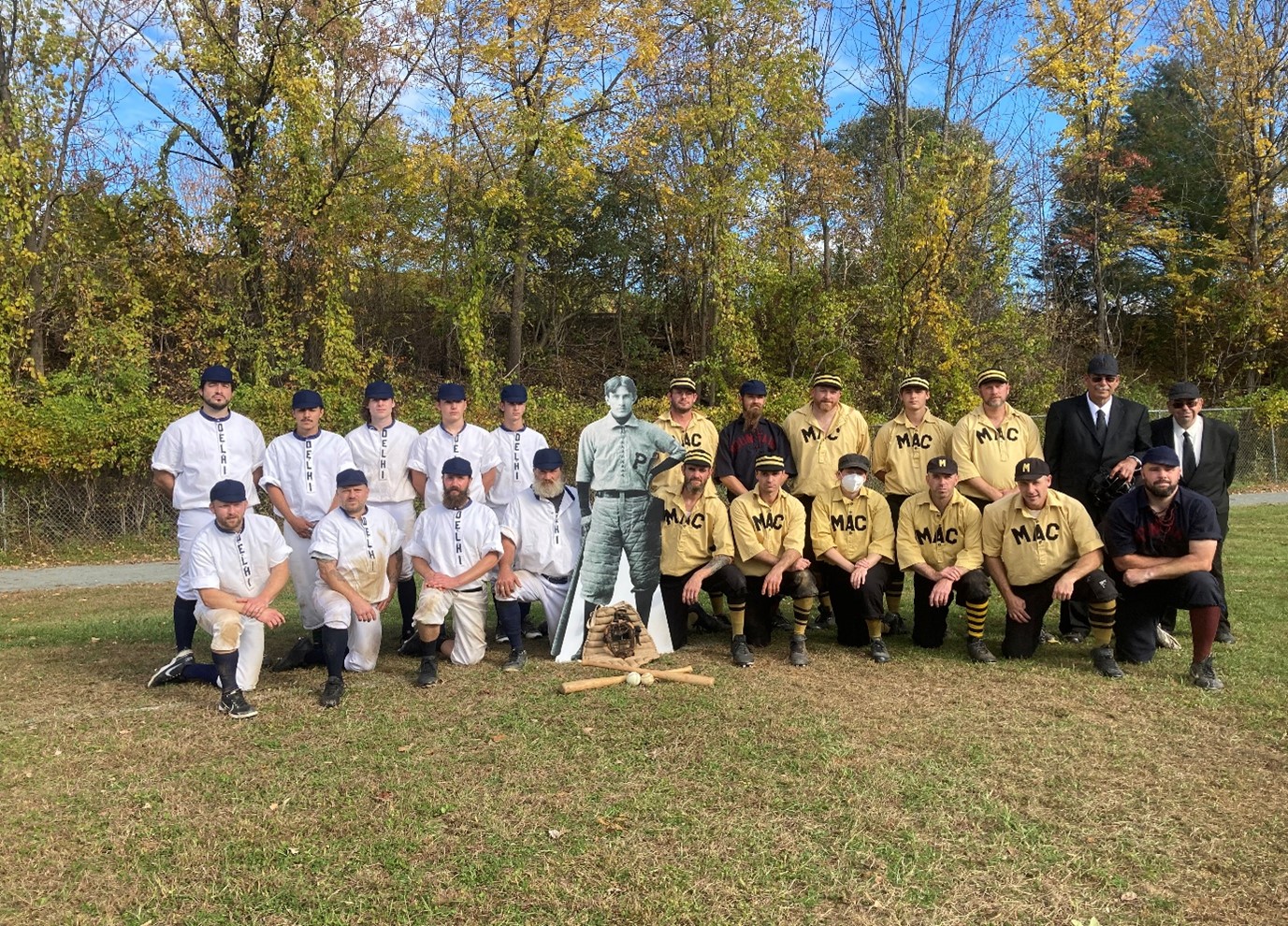 Two baseball teams in vintage baseball uniforms in a baseball field. Green trees in the background.