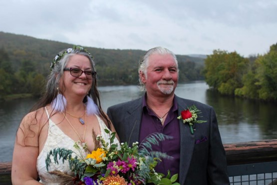 woman wearing glasses, flowered headband, and white lacy dress while holding a bouquet stands with a man in a dark purple button down shirt and navy coat. Both smile against backdrop of Delaware River.
