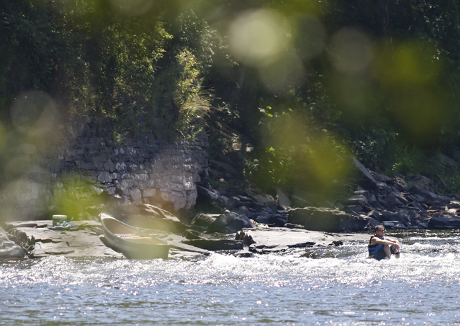 Visitor in life jacket sits in shallow water along river. His boat is bached on some nearby rocks.