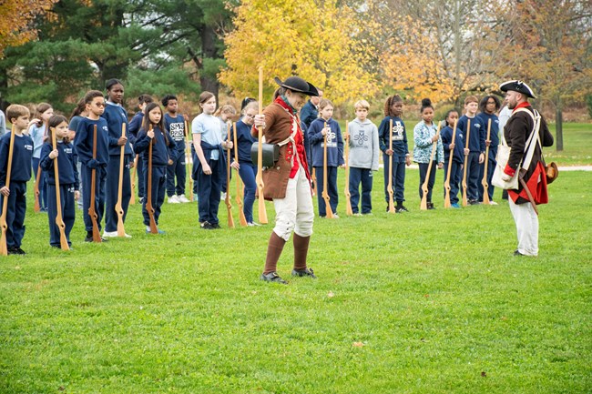 A park ranger wearing a revolutionary war soldier uniform stands in front of a large group of children holding wooden muskets.