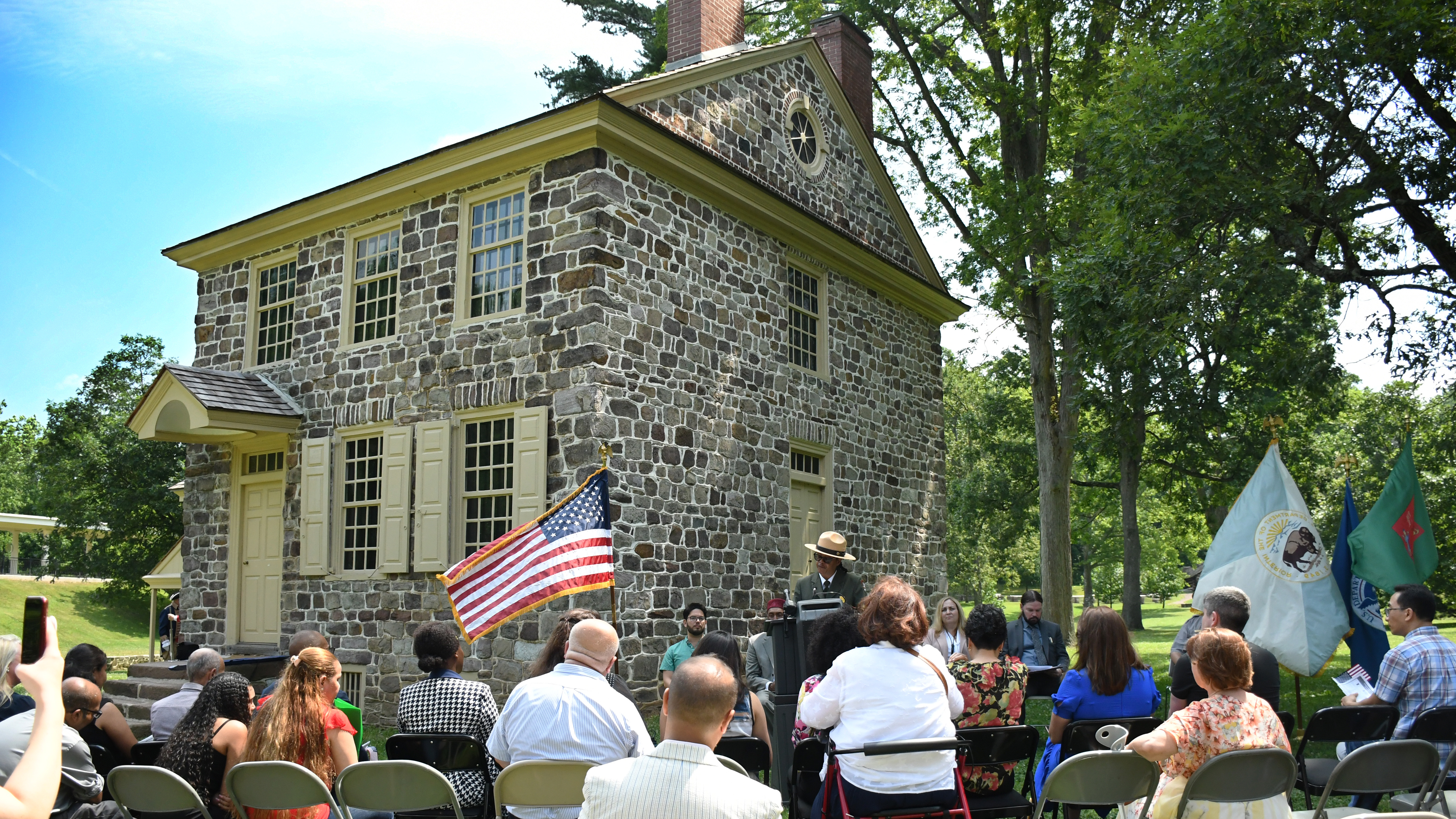 a park ranger at a podium next to a small stone farmhouse speaks to people seated in front