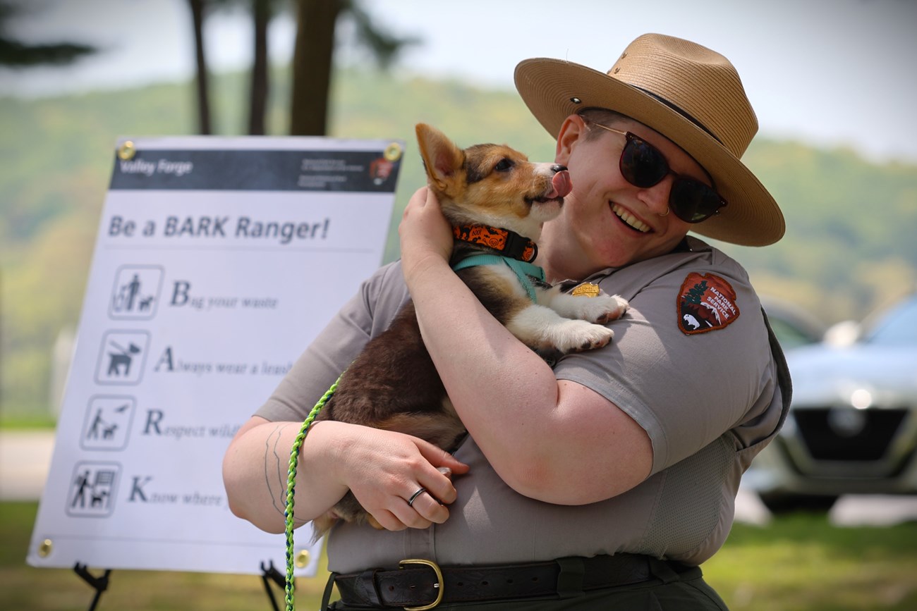 A smiling park ranger holds a small dog in their arms.