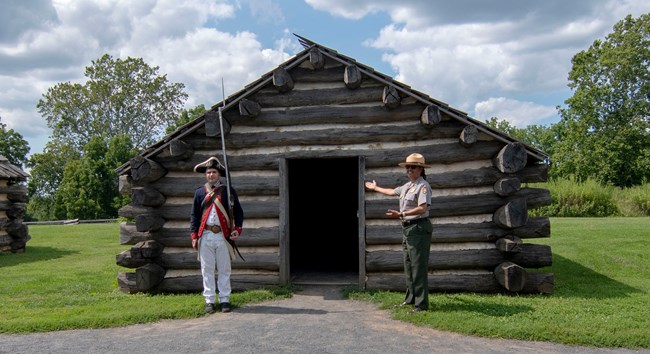 a man wearing a continental army soldier uniform stands next to a uniformed park ranger and a log hut