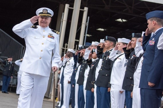 A white uniformed man salutes with other uniformed military personnel alongside
