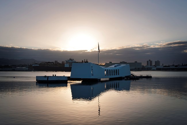 Sunrise view of the USS Arizona Memorial at Pearl Harbor, showing the white structure floating on calm water with the sun rising behind clouds in the background.