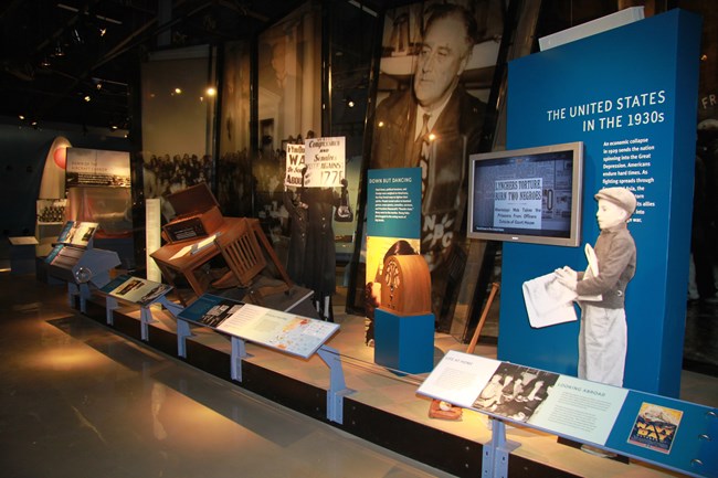 An exhibit at the Pearl Harbor Visitor Center showcasing the United States in the 1930s. The display features life-size cutouts, vintage radios, and informational panels. A figure of a young boy in period attire stands in the foreground.