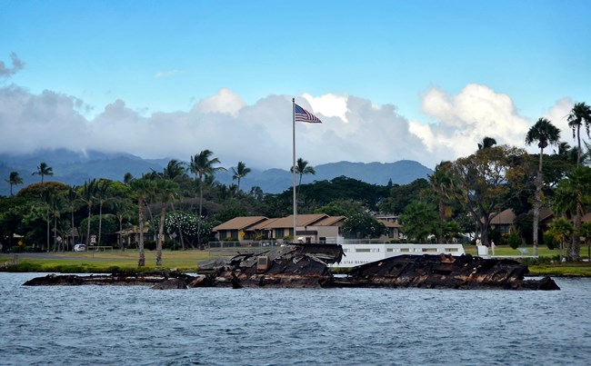 A photograph showing the USS Utah Memorial at Pearl Harbor with remains of the sunken ship visible in the foreground. In the background, an American flag flies atop a pole, with lush green mountains and palm trees under a partly cloudy sky.