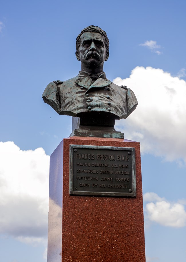 A bronze sculptued figure of a man in a uniform sits atop a pedestal with an inscription on the base.