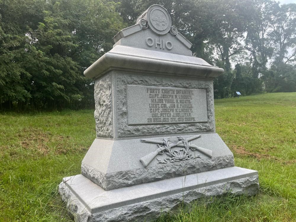 Stone Monument topped by state seal, 2 knapsacks, and 2 crossed rifles with a laurel wreath.