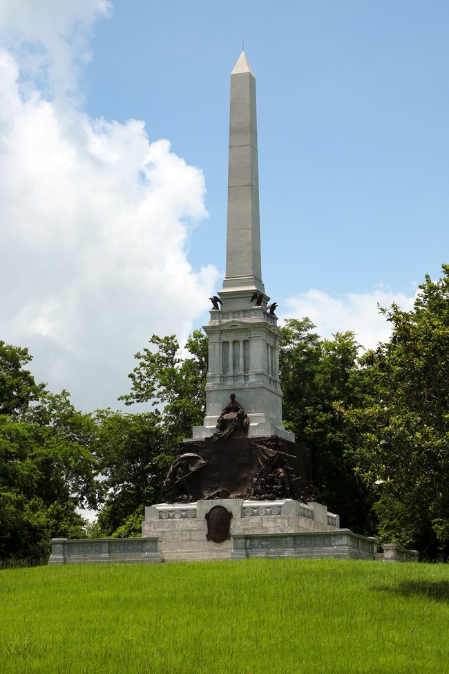 A tall column stands in the middle of a large pedestal. Sitting atop the pedestal is a bronze statue of a woman sitting.
