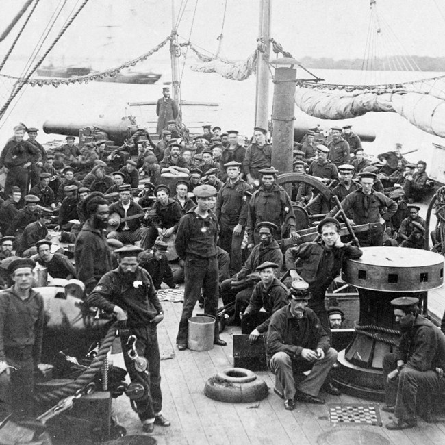 Black and white image of Civil War sailors on the deck of ship. Sailors are standing around, leading on cannons, and playing checkers.