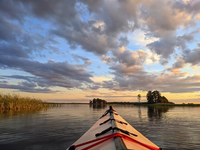 The nose of a kayak points towards a blue and yellow sunset reflected by the waters of a scenic lake.