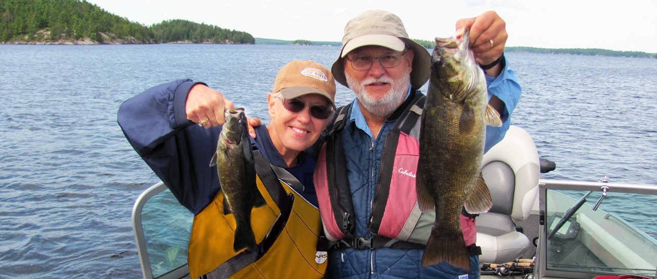 A retirement-aged couple float in a boat on a scenic lake, holding up two Smallmouth Bass fish they recently caught.