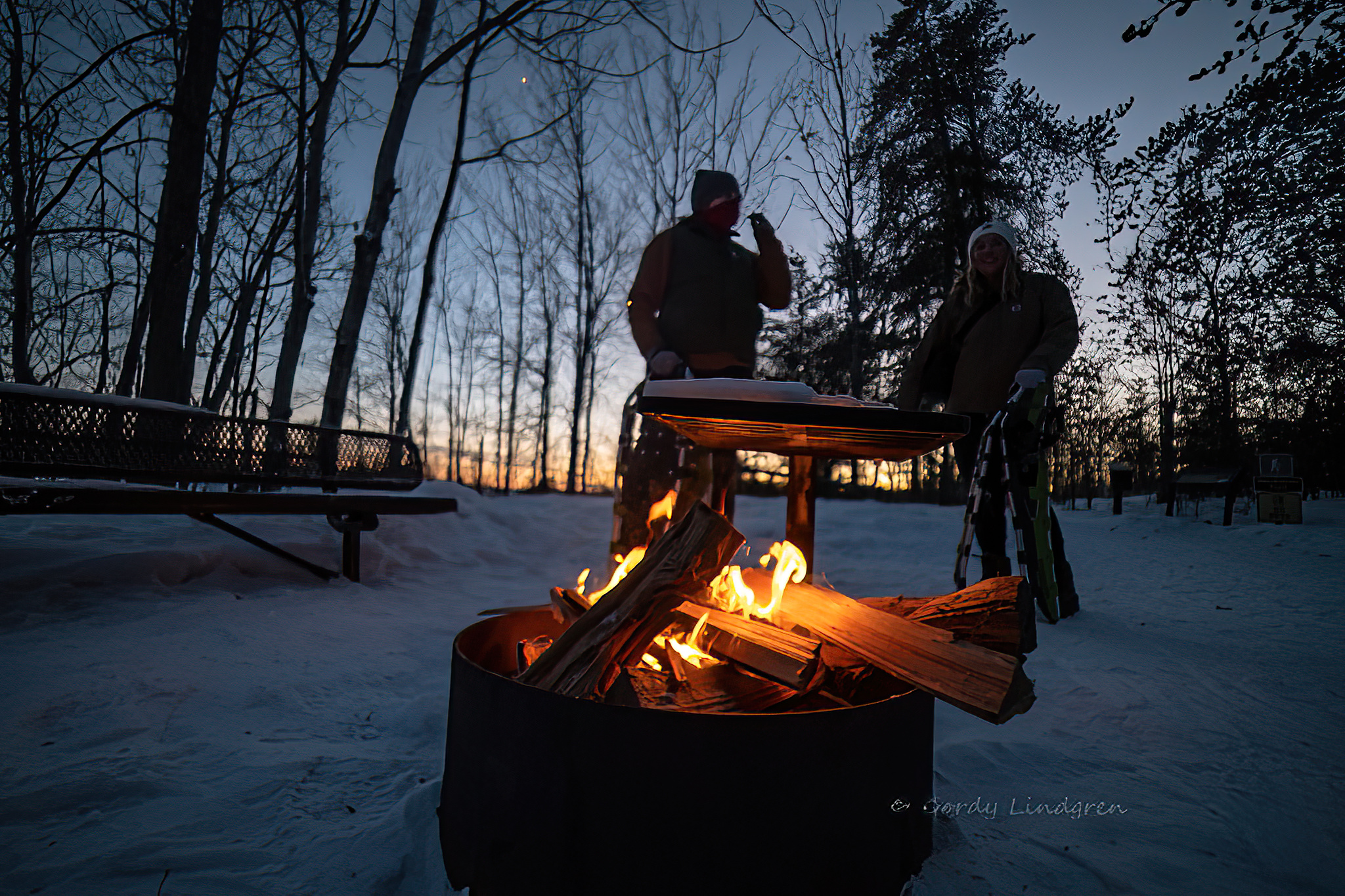 two people with snowshoes gather around a campfire on a snowy evening.