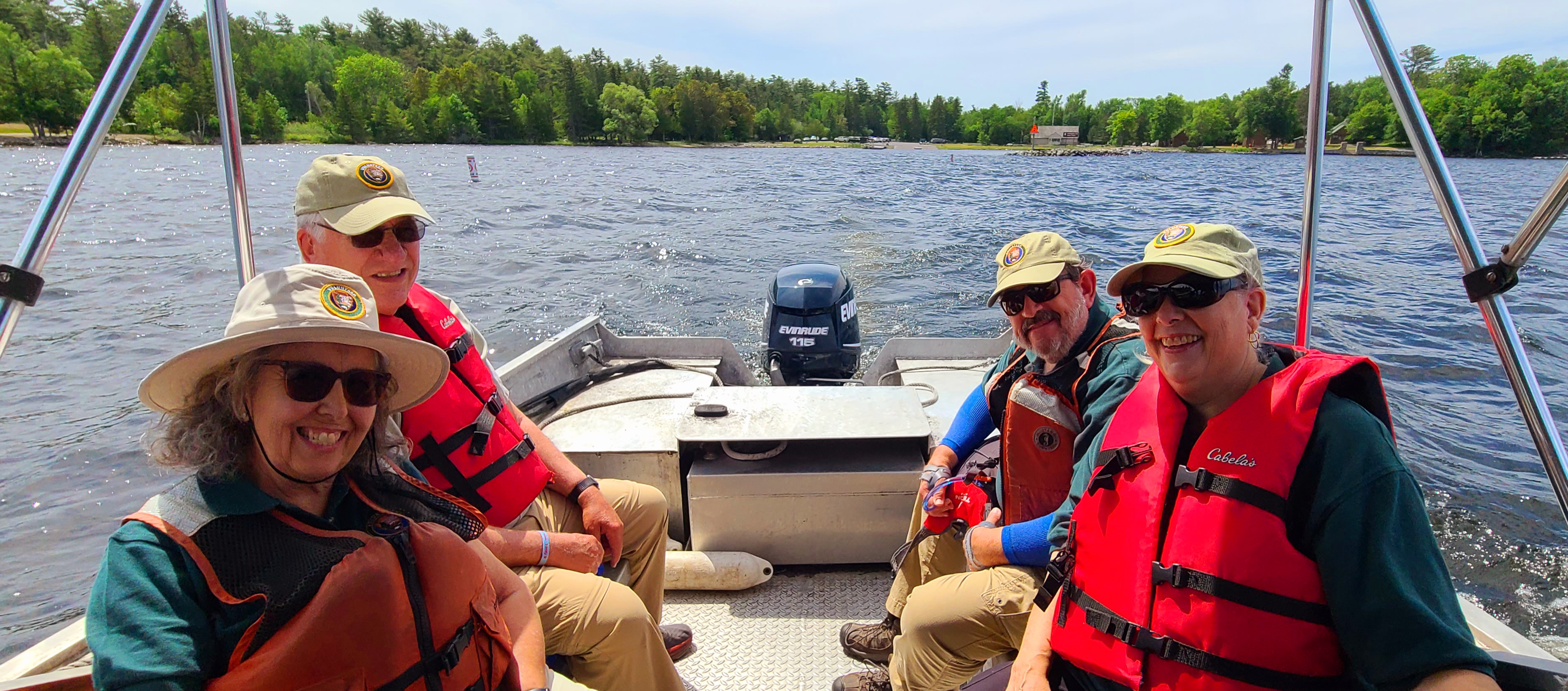 Four boaters wearing U.S. Coast Guard approved orange PFD life jackets smiling while riding on the water with Kabetogama Lake Visitor Center and harbor in the background.