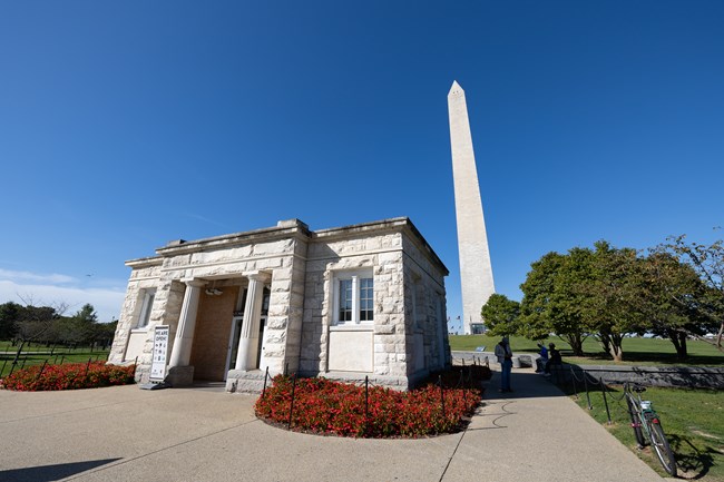A small stone building ringed with red flowers stands below the Washington Monument.
