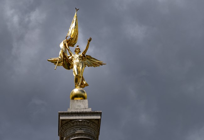 WHHO_Monuments_20250515_NKing_021_crop A statue of a golden, winged person holding her hands aloft with a flag in one, and the other hand open.