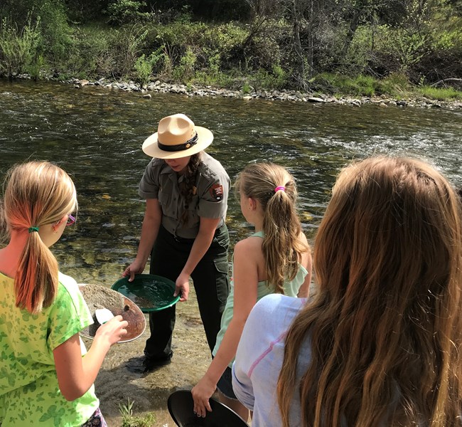 Park ranger with gold pan demonstrating gold panning to students. Clear Creek in background.