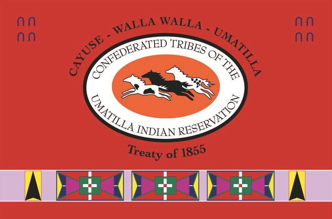 Three horses in an oval with text reading Confederated Tribes of Umatilla Indian Reservation. Text outside oval reads Cayuse - Walla Walla - Umatilla, Treaty of 1855. A band of geometric patterns runs along the bottom on a field of red.