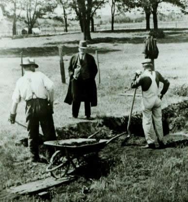 Men with shovels and wheel barrow standing by hole.