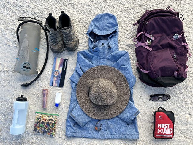 a blue rain jacket, purple backpage, map with White Sands on it, compass, water bottle, water bladder, flashlight, sunscreen bottle, trail mix bag, sunglasses, hiking shoes, and a first aid kit lie on a white sand background.