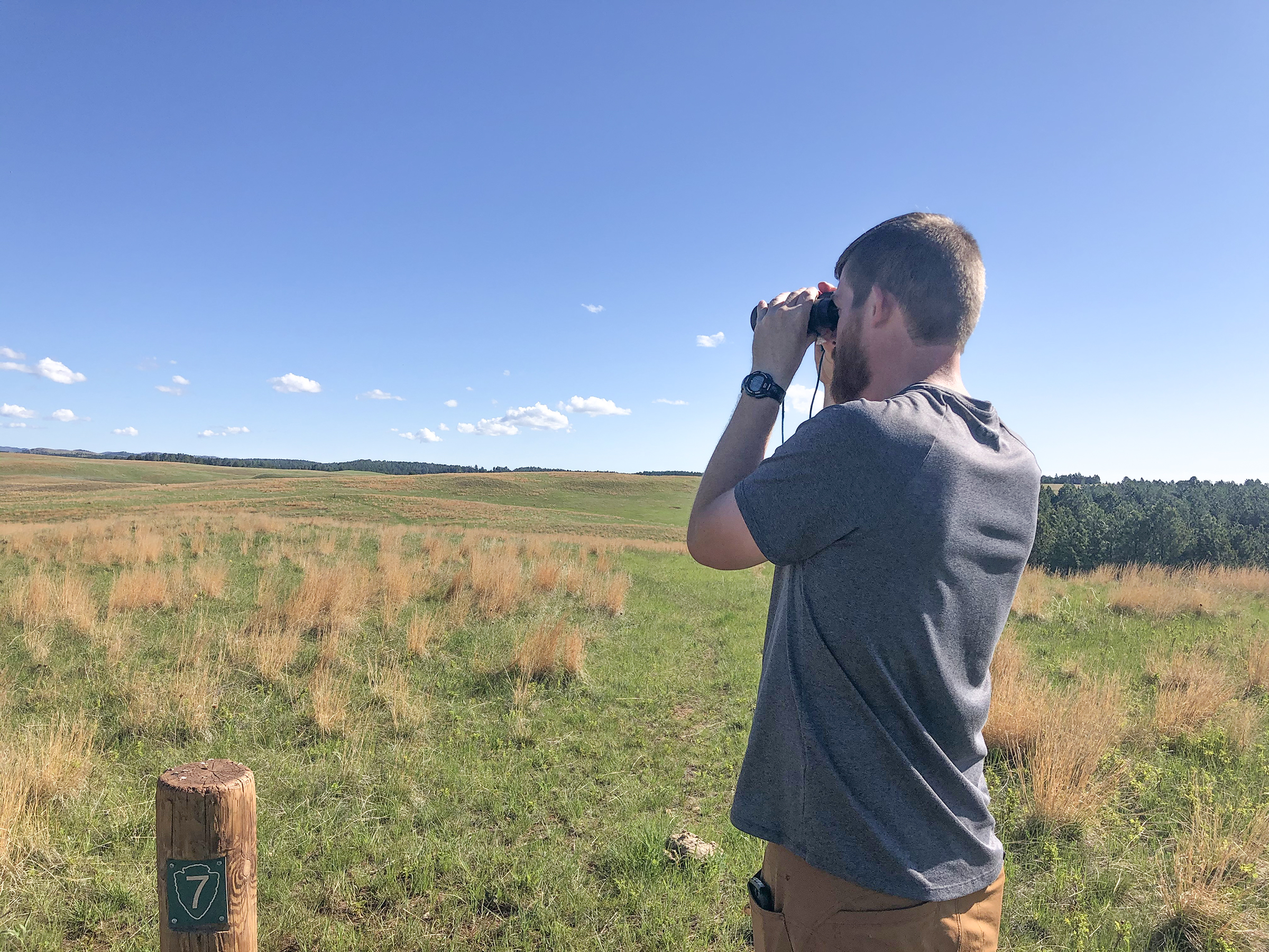 A white male with short hair and bear wearing a gray t-shirt and tan pants looks through binoculars across the prairie under a light blue sky with a few scattered clouds in the distance.