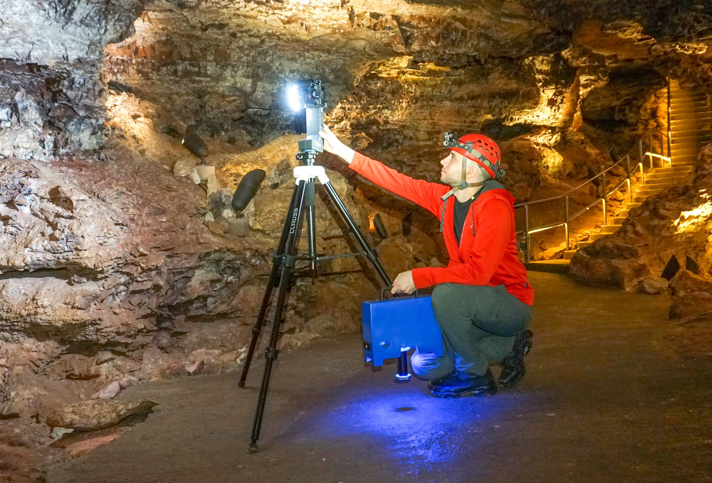 A white male dressed in green pants, red jacket, and red caving helmet kneels on the floor of a cave room while reaching to operated equipment mounted on a tripod.