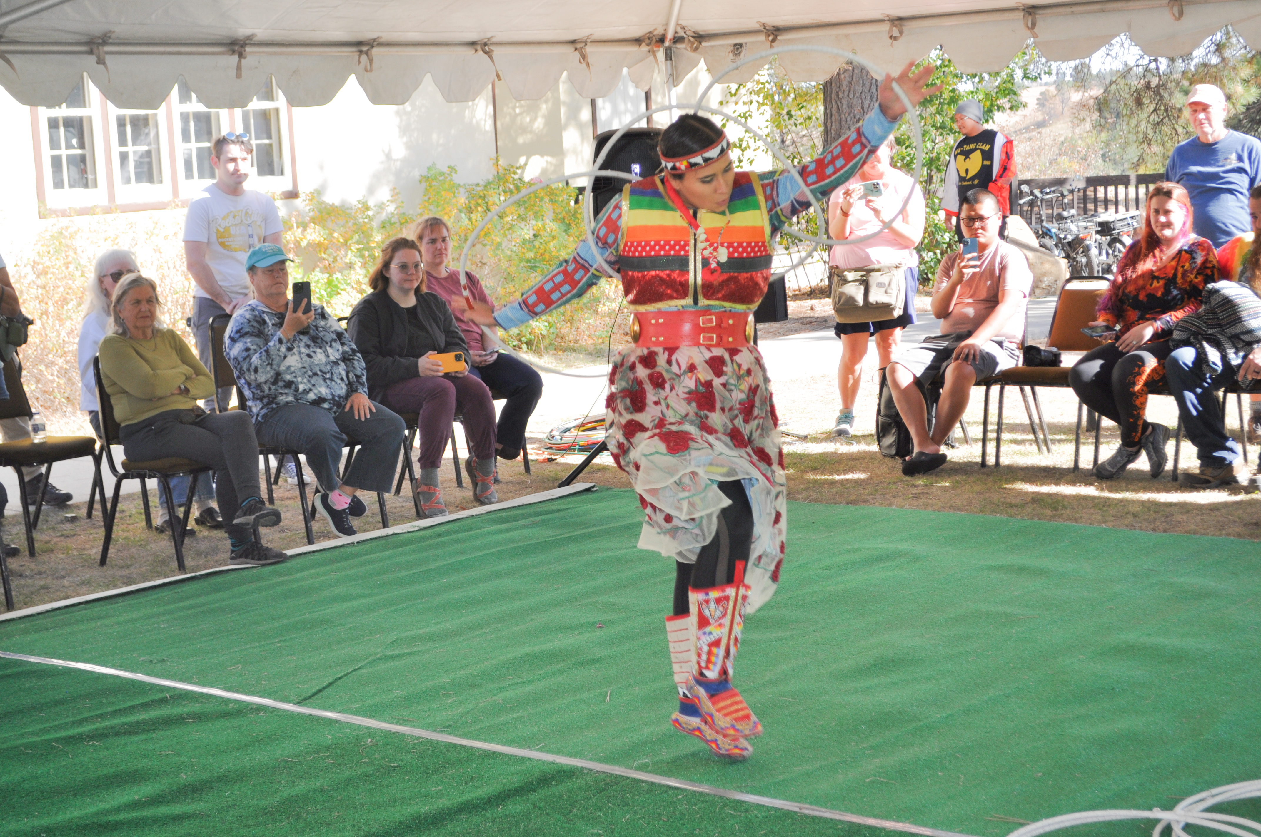 Woman wearing a colorful dress dances on a green carpet with white hoops along her arms and in her hands. People stand and sit along the edges of the carpet watching her.