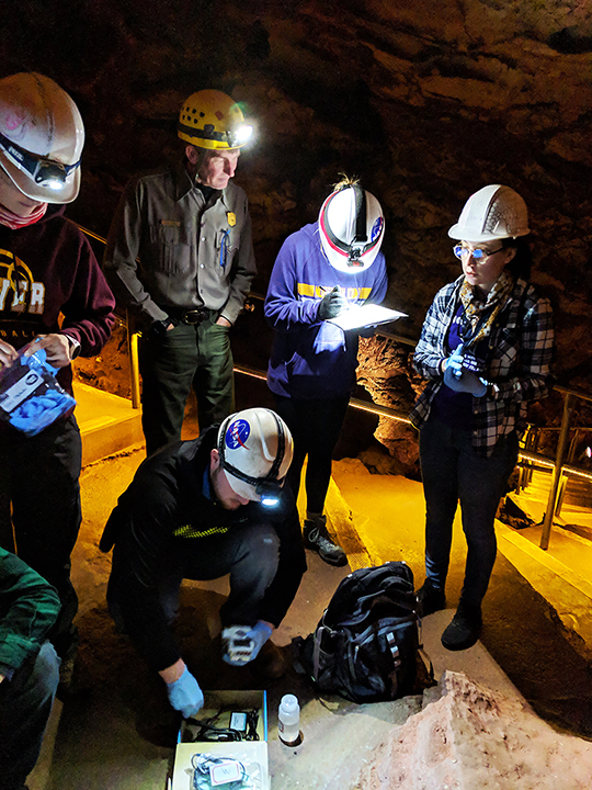 Group of students in the cave, wearing hardhats, gathered in a group as they collect water samples in bottles.