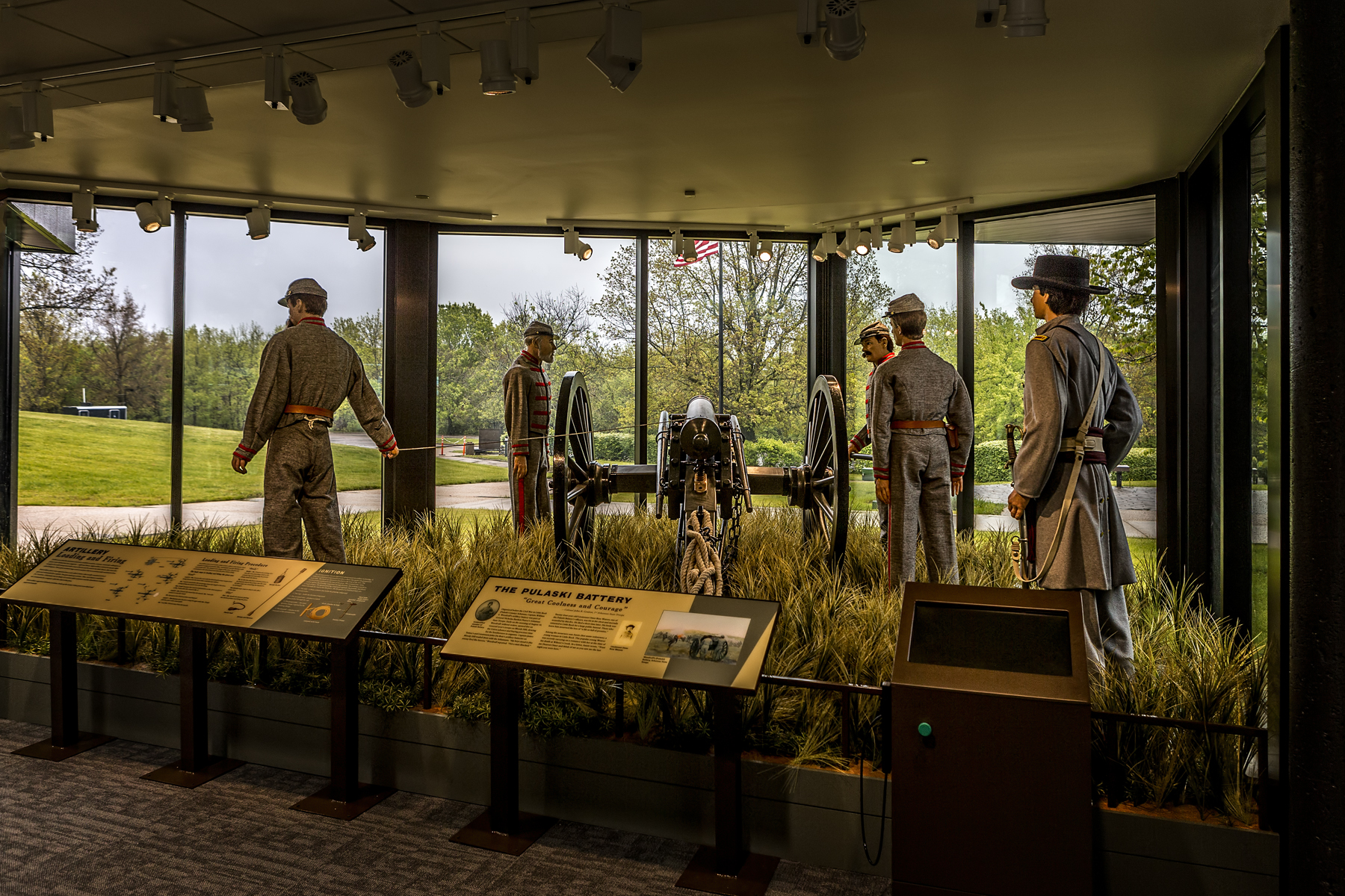 Visitor Center window display featuring a grassy field with 5 cannoneer mannequins in period dress representing The Pulaski Battery in the position of fire at the cannon.