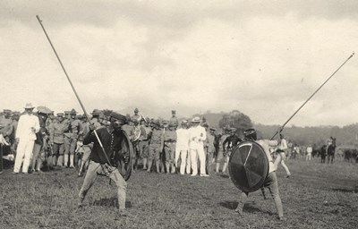 Two indigenous men with spears pointed at each other as spectators look on