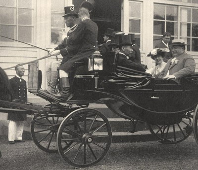 A group of people in a carriage with two men seated up front and one tipping his hat