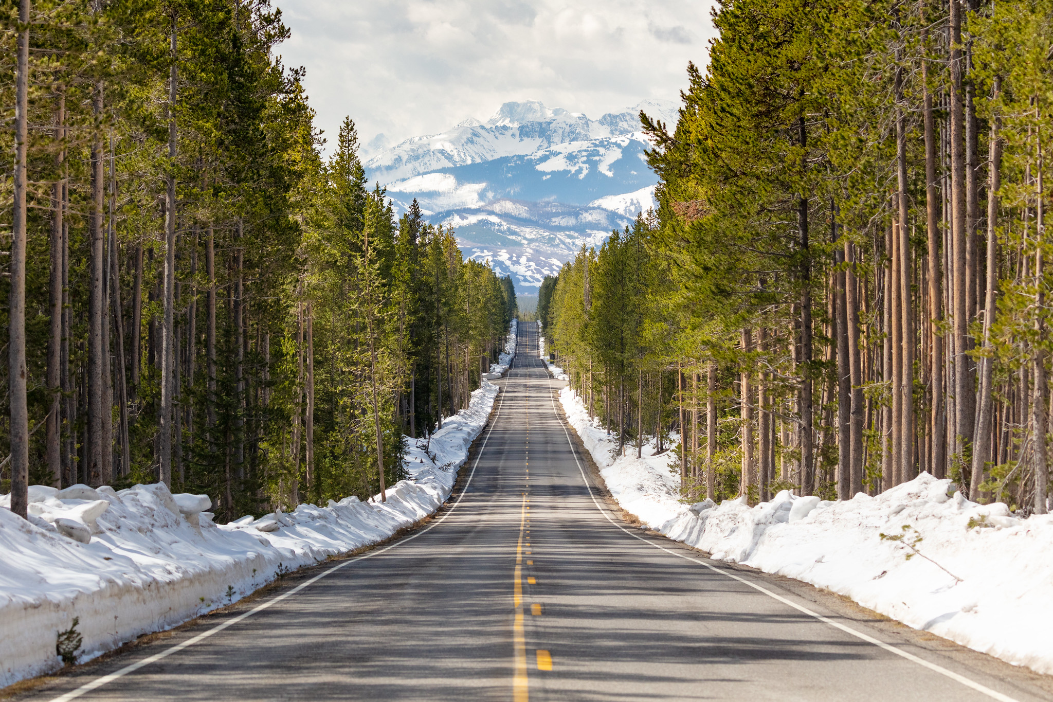 a straight, paved, two-lane road through trees, surrounded by snowbanks and a view of snowy mountains