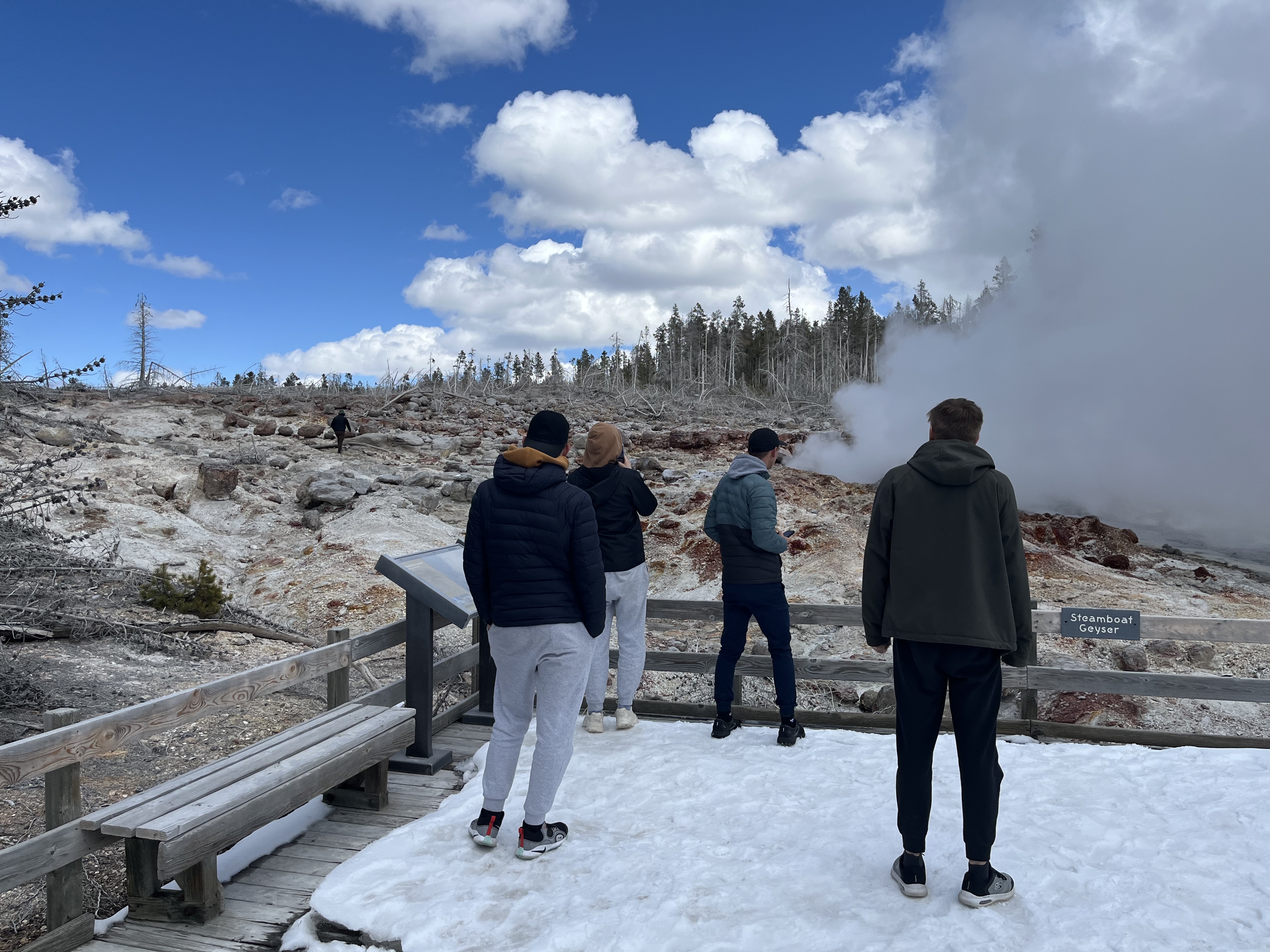 a group of people standing on a boardwalk near a geyser and sign that reads "Steamboat Geyser." Nearby, a person can be seen walking off the boardwalk and onto the geyser feature.