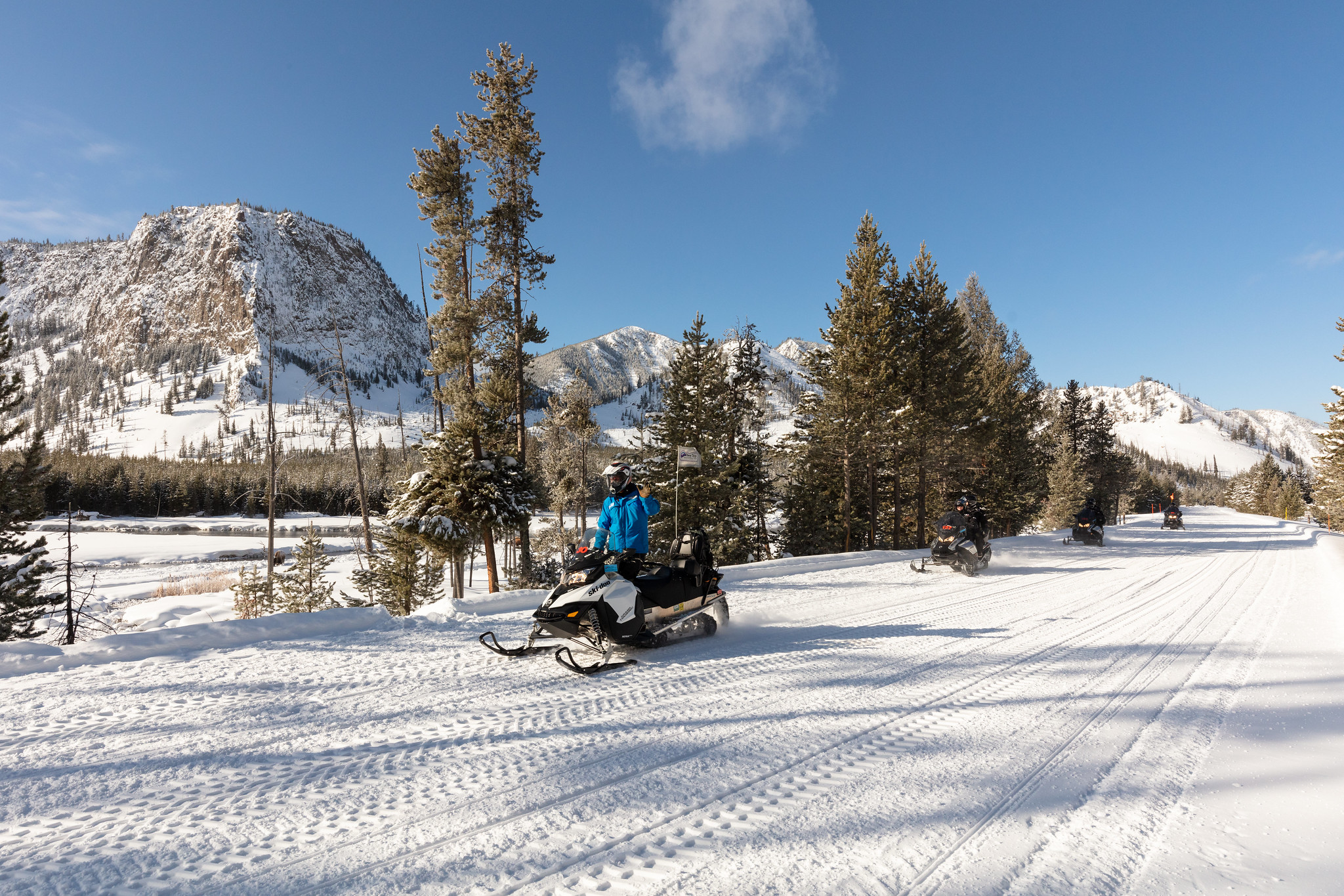 A group of snowmobiles rides on an oversnow road alongside a river.