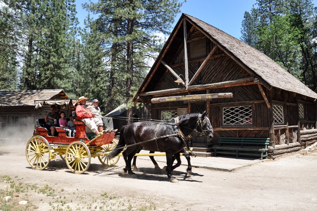 Una carreta tirada por caballos pasa por una cabaña de madera que tiene un letrero que dice: "Compañía de Transporte de Yosemite".