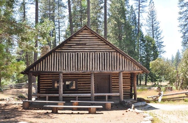 Una cabaña de madera con un porche cubierto enfrente hecho con postes y al fondo se ven varios pinos.