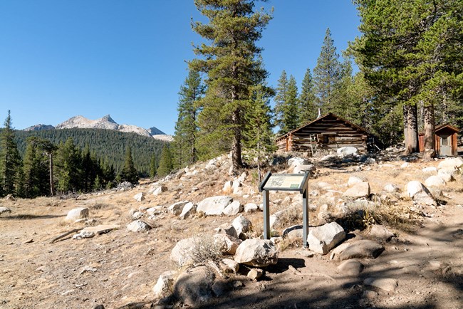 La cabaña de madera se encuentra en la cima de una colina sobre un prado. A lo lejos se pueden ver picos de granito.