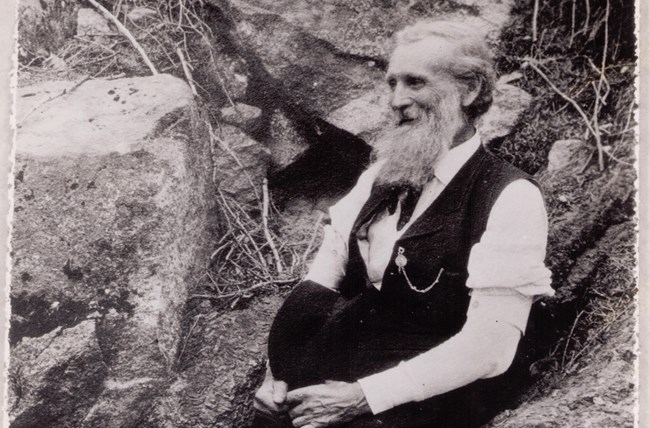 Una foto en blanco y negro muestra a un anciano sentado en una roca. Su cabello blanco y su barba larga.