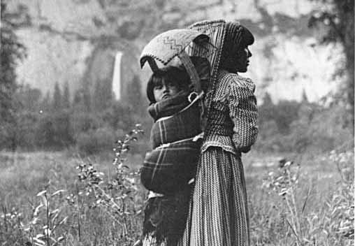 Un bebé está envuelto en una manta y asegurado a una canasta como cuna que usa en la espalda la nativa que está de pie en el prado. Se ve una cascada a lo lejos.