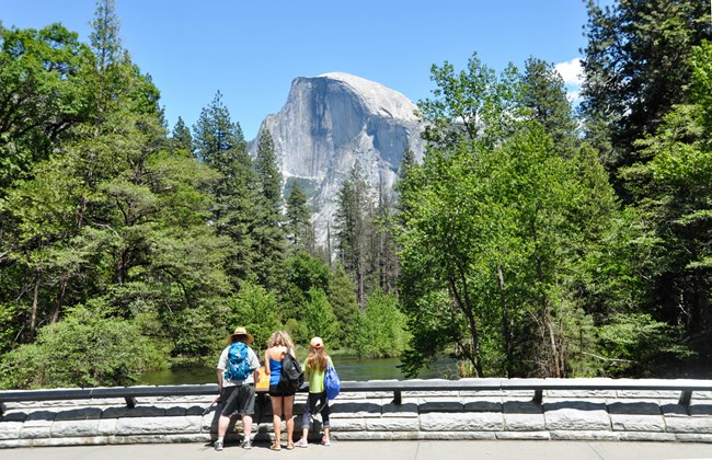Tres personas se detienen y miran un cartel al costado del camino que está unido a la barandilla del puente. Más allá del mismo, el Half-Dome se eleva por encima de la línea de árboles.
