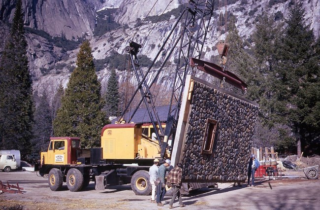 Construction equipment hauls a large panel of rock wall into place with workers nearby