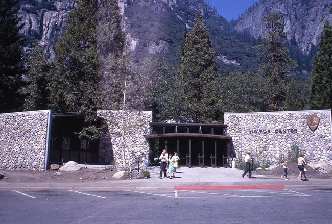 A wide, low building with rock walls and a glass-paneled entryway; visitors stand and walk near the building