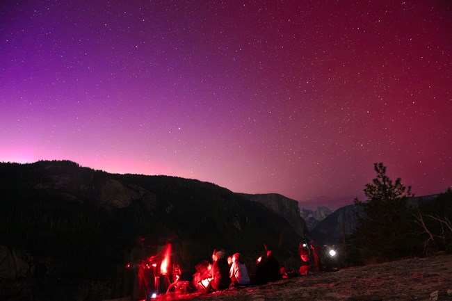 A group of people sit on granite, surrounded by the glow of red light and looking up at a starry sky.