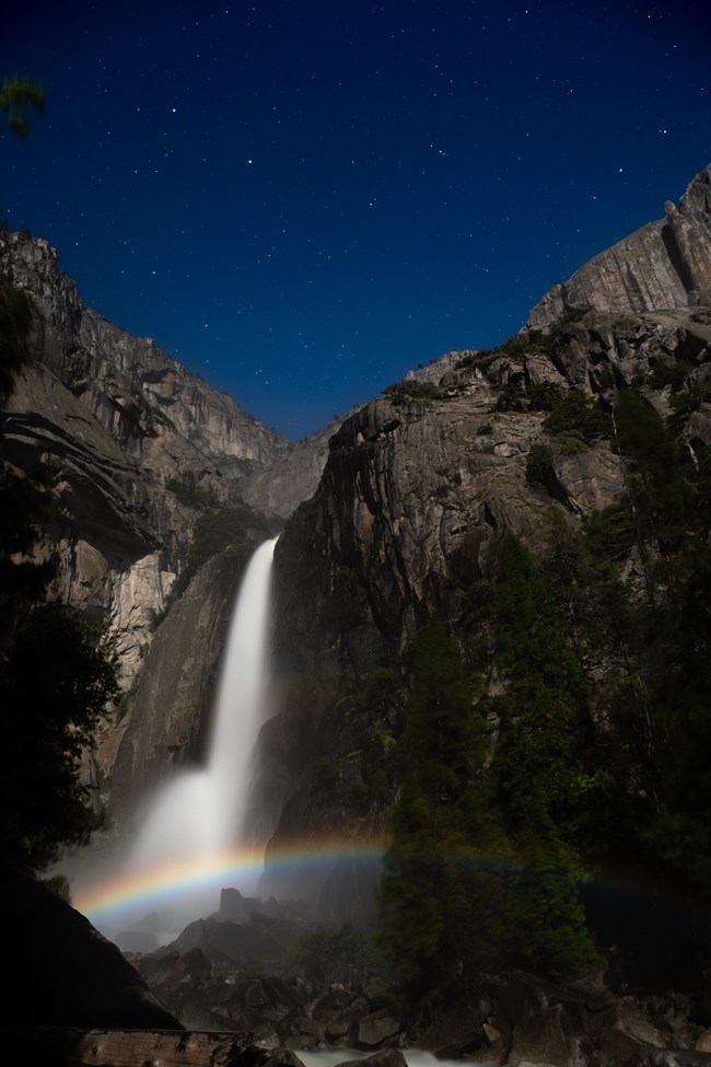 A waterfall cascades below a starry blue sky, with a rainbow in the foreground.