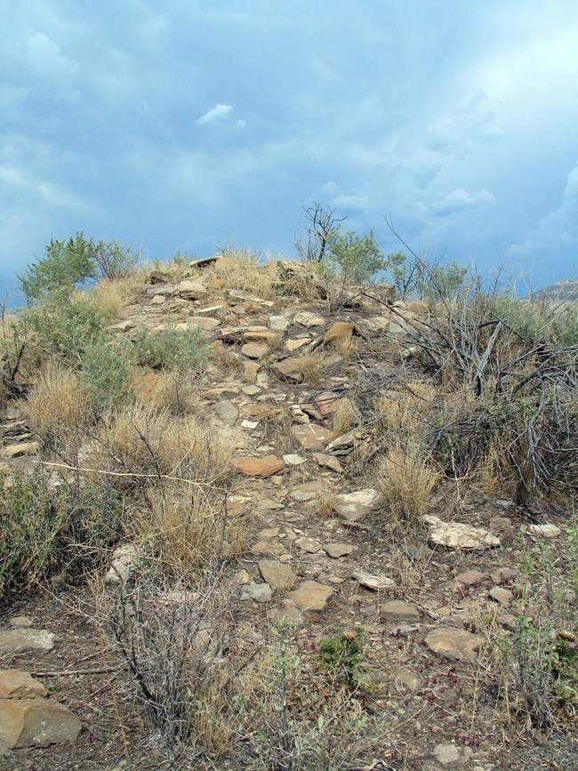 A hill with rocks and stones which are mostly flat, ranging in color from white to muted orange, surrounded by green and brown shrubs. A blue and white sky