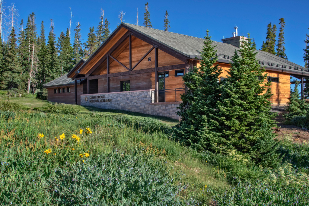 A wood and metal building surrounded by green trees and grass. A rock wall in front of the building reads cedar breaks national monument with an arrowhead next to it