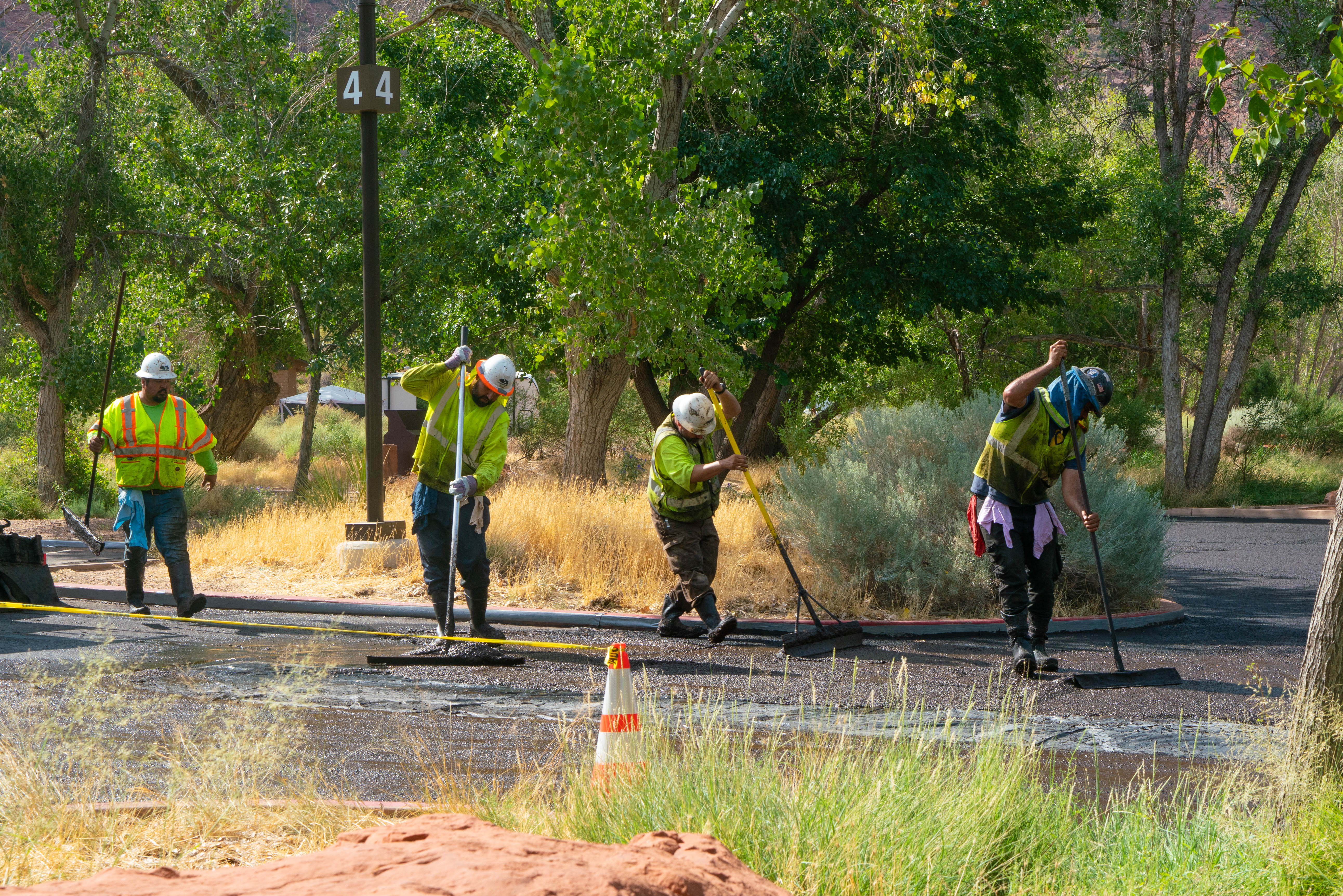 Four men in hard hats and yellow vests push black material onto a road