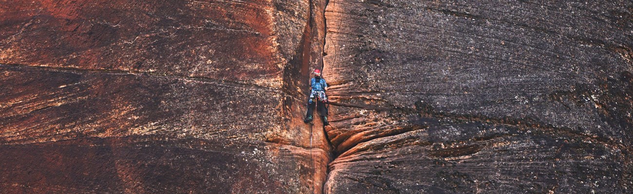 A climber with a red helmet and blue shirt ascends a cliff face in Zion National Park.